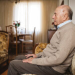 A senior man sits on a sofa in the living room, facing sideways.
